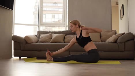 Blonde,-caucasian-woman-in-black-leggings-and-sports-bra-doing-youga-or-stretching-body-while-sitting-on-a-training-mat-at-bright-living-room-with-a-big-couch-on-the-background