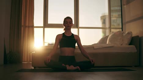 Gorgeous-scene-of-blonde-female-adult-meditating-in-lotus-position-at-home-sitting-on-yoga-mat,-enjoying-exercises.-Young-people,-healthy-lifestyle-and-interiors-concept.-Mild-sunset-on-the-background-window