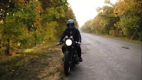 Unrecognizable-man-in-black-helmet-and-leather-jacket-stopping-his-vintage-motorcycle-on-the-road-side-on-sunny-autumn-day