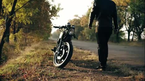 Unrecognizable-man-in-helmet-and-leather-jacket-coming-up-to-his-bike-and-starting-the-engine-while-standing-on-the-roadside-in-a-sunny-day-in-autumn