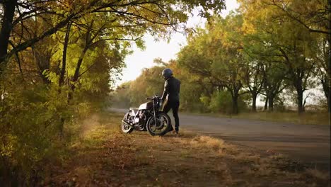 Unrecognizable-stylish-man-in-helmet-and-leather-jacket-coming-up-to-his-bike-and-starting-the-engine-while-standing-on-the-roadside-in-a-sunny-day-in-autumn