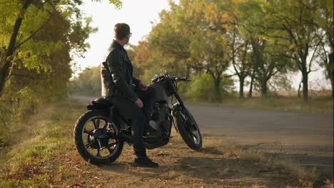 Young-attractive-man-in-leather-jacket-and-stylish-sunglasses-is-sitting-on-his-bike-on-the-country-road-on-a-sunny-day-in-autumn