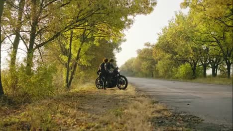 Young-attractive-couple-starting-to-ride-a-motorcycle,-traveling-together-on-the-asphalt-road-on-a-sunny-day-in-forest-in-autumn