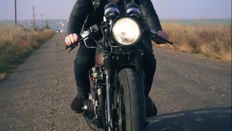 Front-view-of-a-unrecognizable-man-in-black-leather-jacket-riding-motorcycle-on-a-country-road-holding-a-handlebar.-Big-headlight-turned-on