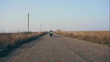 Stylish-cool-young-man-in-sunglasses-and-leather-jacket-approaching-while-riding-a-motorcycle-on-a-road-on-a-sunny-day.-Then-he-outruns-the-camera-point-of-view-and-moves-forward.-Shot-in-4k