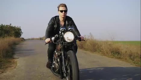 Portrait-of-a-stylish-cool-young-man-in-sunglasses-and-leather-jacket-riding-motorcycle-on-a-road-on-a-sunny-day