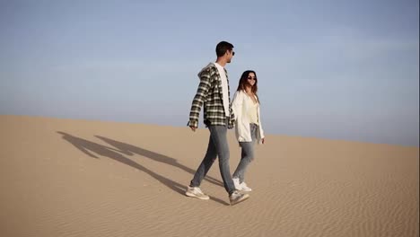 Happy-couple-holding-hands-walking-in-romantic-relationship-under-sun-and-blue-sky-in-desert.-Two-young-lovers-walking-by-desert-sand-in-casual-clothes-and-smiling