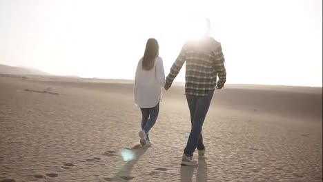 Rare-view-of-couple-holding-hands-walking-in-romantic-relationship-under-sun-and-blue-sky-in-desert.-Two-young-lovers-walking-by-desert-sand-in-casual-clothes,-holding-hands.-Lens-flares