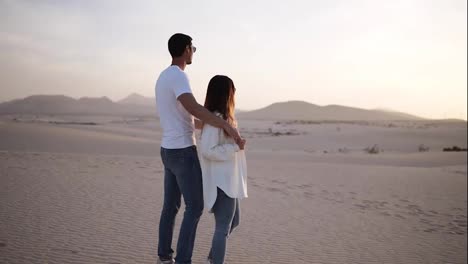 Young-couple-standing-and-hugging,-man-holding-her-woman-from-the-back-on-the-dried,-sandy-desert-watching-the-landscape-together-thoughtfully