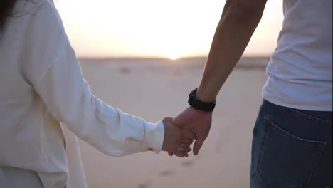 Aimed,-close-up-footage-of-holding-hands-couple-walking-in-romantic-relationship-under-sun-and-blue-sky-in-desert.-Two-young-lovers-walking-by-desert-sand-in-casual-clothes.-Landscape-on-background