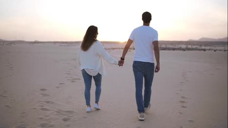 Backside-footage-of-holding-hands-couple-walking-in-romantic-relationship-under-sun-and-blue-sky-in-desert.-Two-young-lovers-walking-by-desert-sand-in-casual-clothes.-Landscape-on-background.-Slow-motion