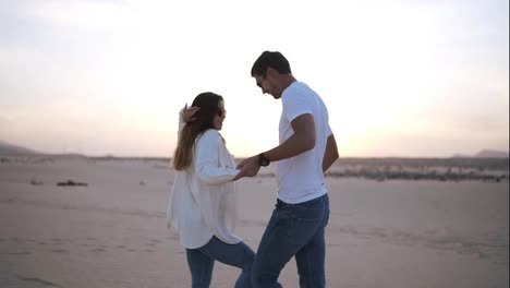 Nice-lovely-cheerful-laughing-couple-wearing-casual-white-T-shirts.-Dark-haired-guy-spinning-with-his-lady,-holding-hands-in-moves.-Dancing-on-nature-in-empty-desert