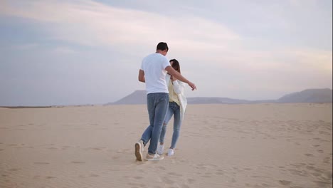 Nice-lovely-cheerful-laughing-couple-wearing-casual-white-T-shirts.-Dark-haired-guy-spinning-with-his-lady,-holding-hands-in-moves.-Dancing-on-nature-in-empty-desert.-Full-length.-Slow-motion