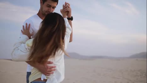 Nice-lovely-cheerful-couple-wearing-casual-white-T-shirts.-Dark-haired-guy-spinning-with-his-lady,-holding-hands-in-moves.-Dancing-on-nature-in-empty-desert.-Sensual-moves.-Slow-motion