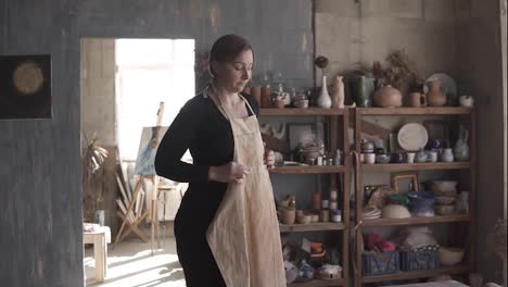 Slow-motion-footage-of-young-caucasian-woman-in-casual-clothes-standing-in-pottery-workshop,-with-ceramic-wares-and-supplies-on-shelves-and-table-with-tools-in-middle,-and-putting-on-beige-apron