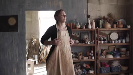 Slow-motion-footage-of-young-caucasian-woman-in-casual-clothes-in-pottery-workshop,-with-ceramic-wares-and-supplies-on-shelves-and-table-with-tools,-putting-on-beige-apron-and-smiling
