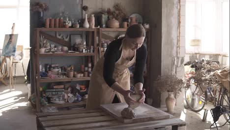 Portrait-of-female-potter-wearing-beige-apron-putting-clay-piece-on-worktop-and-then-starting-kneading-it-with-her-hands.-Pottery-products-on-shelf-behind