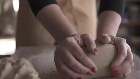 Close-up-footage-of-female-hands-with-beautiful-red-manicure-holding-clay-and-kneading-it-on-a-worktop.-Wearing-beige-apron.-Unrecognizable
