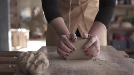 Close-up-footage-of-female-hands-with-beautiful-red-manicure-holding-clay-and-kneading-it-on-a-worktop.-Wearing-beige-apron.-Unrecognizable-person.-Overview.-Blurred-background
