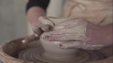 Close-up-of-potter's-dirty-hands-working-with-wet-clay-on-a-pottery-wheel-making-a-vase-in-a-workshop.-Unrecognizable-female-person-forming-product