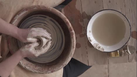 Close-up-top-view-of-potter's-hands-with-working-with-wet-clay-on-a-pottery-wheel-making-a-clay-product-in-a-workshop.-Unrecognizable-female-person-formong-vase,-bowl-with-a-water-on-a-table