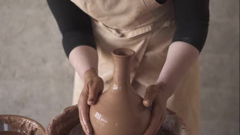 Cheerful,-young-good-looking-woman-wearing-apron-sitting-in-pottery-and-making-clay-jug-on-potter-wheel-using-hands.-Woman-smiling-to-a-camera