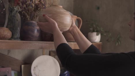 Side-view-portrait-of-contemporary-female-potter-putting-handmade-vase-on-shelf-in-studio,-pleased-with-her-work,-smiling-to-the-camera.-Slow-motion