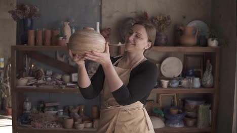 Light-brown-vase-from-clay-in-the-female-potter's-hands.-A-young-and-cheerful-woman-holding-a-vase-of-clay.-The-potter-works-in-a-pottery-workshop-with-clay.-Enjoying-the-perfect-shape