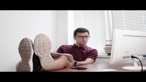 Young-worker-in-glasses-holding-the-keyboard-on-his-legs-typing-and-looking-at-the-screen-of-the-computer-in-office-with-his-legs-on-the-table.-Busy-tired-worker-in-the-modern-office.-4k