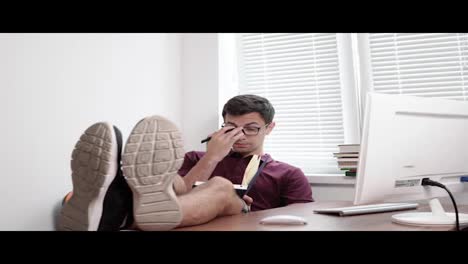 Young-office-worker-in-glasses-holding-the-notebook-in-his-hands-and-making-notes-with-his-legs-on-the-table.-Busy-tired-worker-in-the-modern-office.-Shot-in-4k