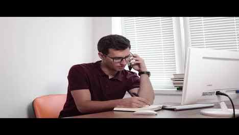 Concentrated-young-businessman-in-glasses-writing-notes-while-talking-on-mobile-phone-with-client-at-the-office.-Shot-in-4k