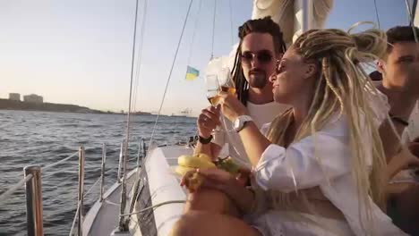 Stylish-couple-with-dreadlocks-in-white-clothes-and-sunglasses-sitting-embracing-on-the-edge-of-the-yacht-smiling,-clinking-with-champangne-glasses.-Loving-couple-spending-time-together-on-the-yacht-beyond-the-sea
