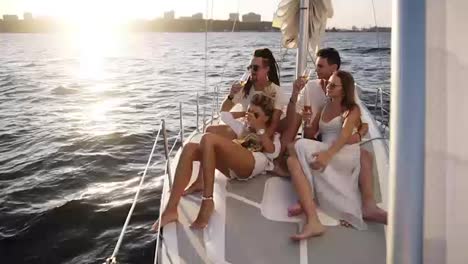 Gorgeous-scene-of-company-of-young-happy-friends-sail-on-a-yacht-bow-on-a-summer-day,-friends,-men-and-women-relax-in-the-sun.-Beautiful,-mild-sunlight-and-urban-silhuettes-on-the-background