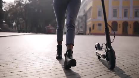 Attractive,-confident-afro-american-girl-in-stylish-sunglasses-and-blue-jeans-walking-carefree-by-morning-city-street-with-her-black-electric-scooter.-Front-view.-Slow-motion