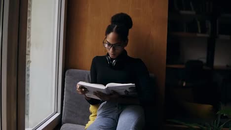 The-young-attractive-afro-american-woman-is-reading-the-book-on-the-wide-window-sill.-Close-up-portrait-of-a-stylish-girl-in-eyeglasses-and-headphones-reading-or-studying