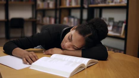 A-female-afro-american-student-in-black-sweater,-tired-and-fell-asleep-on-a-table-with-books-and-notes-in-the-modern-library.-Education,-people,-student-and-learning-concept.-Front-view