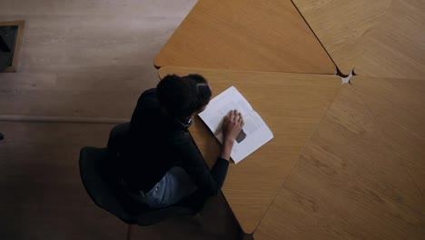 Top-view-footage-of-concentrated-african-young-woman-sitting-by-the-round-wooden-desk-and-reading-book,-studying-for-university-in-the-modern-library,-preparing-for-exams.-Wearing-black-sweater