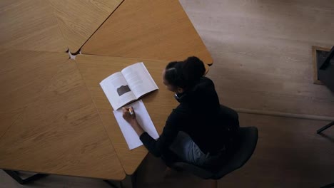 Attractive-concentrated-african-young-woman-sitting-by-the-desk-and-making-some-notes-in-her-note-book-for-university-in-the-modern-library,-preparing-for-exams.-Wearing-black-sweater-and-headphones.-Top-view-footage
