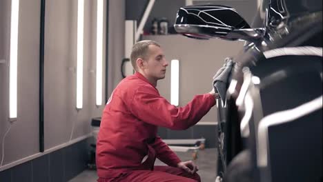 Man-in-red-worksuit-rubs-black-expesive-car-with-microfiber-wiper.-Polishing-at-autocenter.