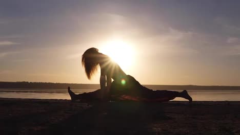 Young-woman-doing-split,-yoga-posture-outdoors-on-the-beach.-Beautiful-silhouette-of-athlete.-Sunset