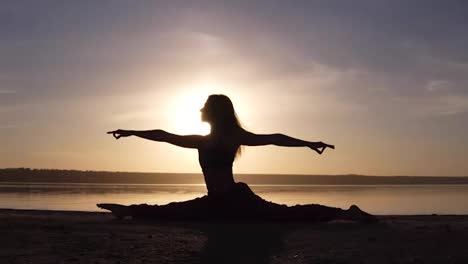 Beautiful-woman's-silhouette-doing-yoga,-split-on-sea-coast.-Morning-sunrise,-smog-sun-on-thee-backstage