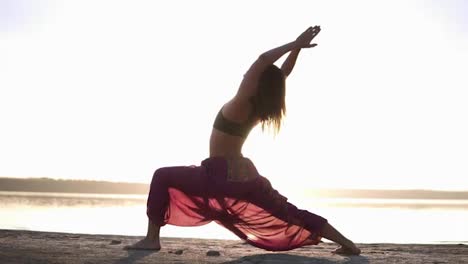 Side-view-of-attractive-young-woman-practicing-yoga-in-warrior-pose-Virabhadrasana-on-seashore.-Light-sun-haze