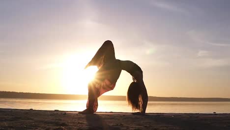 Eine-Junge-Frau-In-Wunderschönen-Farbigen-Yogahosen-Macht-An-Einem-Leeren-Strand-Die-Yoga-Übung-Urdhva-Dhanurasana,-Auch-Bekannt-Als-Aufwärtsbeugenhaltung.-Sonnenlicht-Auf-Dem-Hintergrund