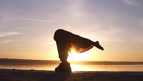 The-girl-does-asana-in-yoga,-slowly-raises-her-legs-in-the-rack-on-her-head.-Silhouette-of-a-woman-in-the-background-of-the-shining-sun