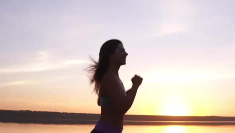 Close-up-footage-of-a-fit-girl-running-along-the-water.-Sunrise.The-young-woman-trainings.-Happy,-smiling