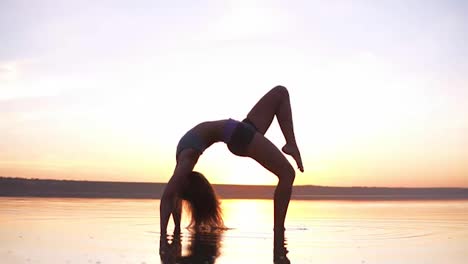 Young-healthy-fitness-woman-doing-yoga,-standing-in-bridge-pose-Setu-Bandhasana-on-the-seaside-in-the-water-at-sunset.-Enjoyment,-harmony-concept