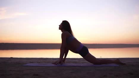 A-sports-brunette-doing-stretching-exercises-on-a-mat-on-the-beach-or-a-lake.-Accelerated-footage.-Front-view