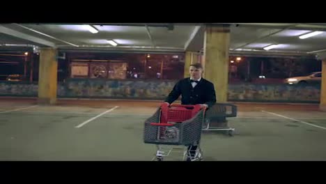 Young-man-in-a-suit-with-bow-tie-moving-shopping-cart-to-his-black-car-in-the-parking-at-night