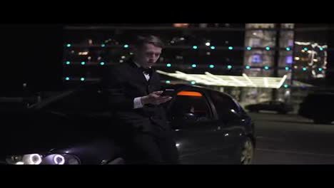 Businessman-in-a-suit-using-a-smartphone-next-to-an-luxurious-executive-car-and-a-big-window-at-the-parking.-Lights-at-the-background