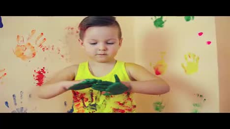 Close-up-of-little-cute-child-with-painted-in-green-hands-making-colorful-handprints-on-the-wall-behind-him.-Playing-and-having-fun.